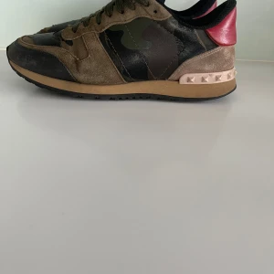 Valentino Rockrunner i camouflage - Snygga Valentino Rockrunner sneakers i en cool kombination av camouflage och rött. Skorna har en unik design med nitar på hälen och en mix av mocka och skinnmaterial. Perfekta för att outfit en unik touch. Skick 8/10🌟Tveka inte på att ställa frågor vid minsta lilla fundering!