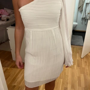Vit plisserad klänning - Snygg vit klänning med en axel och plisserad design från Dry Lake. Den har en oneshoulder-stil och är kort i längden. Perfekt för en stilren look. Prislappen är kvar och den är aldrig använd! 