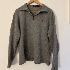 Grå tröja med dragkedja från Ralph Lauren - Säljer en stilren grå tröja från Ralph Lauren med dragkedja och klassisk krage. Tröjan har en broderad röd logotyp på bröstet och långa ärmar. Perfekt för en avslappnad och snygg look.