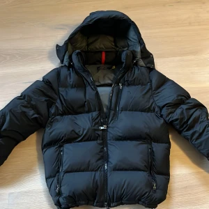 Svart pufferjacka från Polo Ralph Lauren - Säljer en snygg svart pufferjacka från Polo Ralph Lauren. Jackan har en praktisk huva och flera dragkedjefickor. Perfekt för kyliga dagar med sin vadderade design.