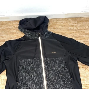 Svart jacka med huva från Boss - Snygg svart jacka från Boss med huva och dragkedja. Jackan har ett unikt mönster på framsidan och är perfekt för en stilren look. Den är långärmad och har en modern design.