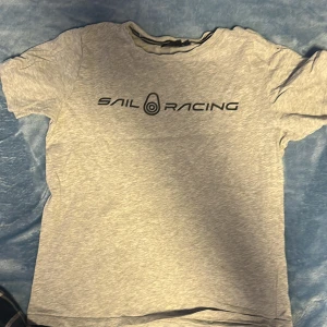 Grå t-shirt från Sail Racing - Säljer en grå t-shirt från Sail Racing med svart logotyptryck på bröstet. T-shirten har en klassisk rund hals och korta ärmar. Perfekt för en avslappnad stil.