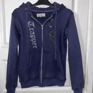Mörkblå hoodie med dragkedja från Trapper - Snygg mörkblå hoodie från Trapper med dragkedja och ett coolt paljettmönster på framsidan. Säljs inte längre, köpt för 900-1000kr. Använd 1 gång pga inte min stil. En schysst tröja som får dig att glänsa lite extra.