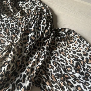 Leopardmönstrad halsduk - Snygg och trendig halsduk med leopardmönster i svart och beige. Perfekt för att ge din outfit en djärv touch. Materialet är lätt och luftigt, vilket gör den idealisk för flera säsonger.