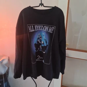 Svart långärmad tröja med 2Pac-tryck - Säljer en svart långärmad tröja med ett stort tryck av 2Pac och texten 'All Eyez On Me' på framsidan. Perfekt för fans av hiphop och streetwear. Tröjan är bekväm och har en normal passform.