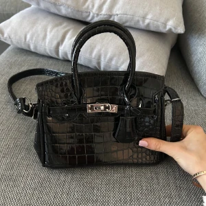 Svart krokodilpräglad handväska - Snygg svart handväska med krokodilpräglat mönster och en elegant metallspänne framtill. Väskan har både handtag och en avtagbar axelrem för flexibilitet. Perfekt för att ge en lyxig touch till din outfit.