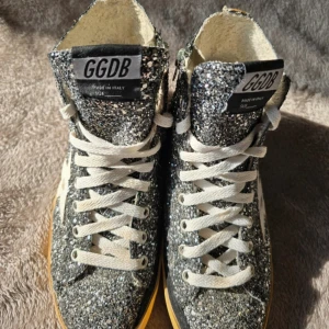 Glittriga sneakers från GGDB - Säljer ett par Golden Goose high-top sneakers i en snygg glittrig design. Skorna är i bra begagnat skick med den karakteristiska vintage-looken som Golden Goose är kända för. Storlek 36.  Saknar tyvärr originalkartong och kvitto därav priset.