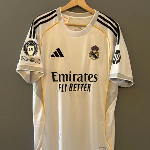 Vit fotbollströja från Adidas 25/26 - Säljer en vit fotbollströja från Adidas med svarta detaljer och klubbmärke på bröstet. Tröjan har korta ärmar och sponsorloggor på framsidan och ärmarna. Nummer och namn på baksidan. Helt ny med tagg en replika i storlek M