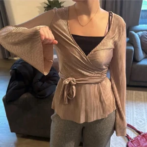 Rosa omlottopp med knytband - Snygg rosa omlottopp med ribbad struktur och knytband i midjan. Toppen har långa ärmar och en v-ringad design som ger en elegant look. Perfekt för att klä upp en vardagsoutfit eller för en kväll ute.