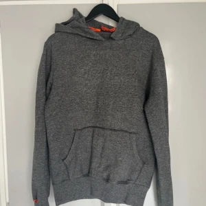 Grå hoodie från Superdry Medium - Säljer en grå hoodie från Superdry med broderad logga på framsidan. Tröjan har en klassisk känguruficka och en bekväm huva. Snören saknas i luvan och slitning i söm vid halsen syns på bild.