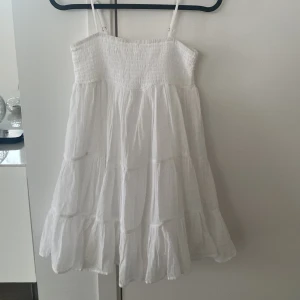 Vit klänning från Gina Tricot - Söt vit klänning från Gina Tricot med smala axelband och smockdetaljer upptill. Klänningen har en luftig och lätt design, perfekt för varma dagar. Den är ärmlös och har en avslappnad passform som ger en skön känsla.