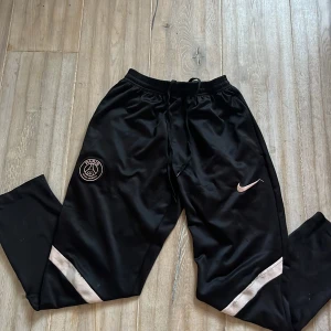 Svarta träningsbyxor från Nike - Snygga svarta träningsbyxor från Nike med PSG-logga på ena benet. Byxorna har en elastisk midja med snörning och vita detaljer vid vaden. Perfekta för sport och träning. Och byxorna har ett litet mini mini hål på framsidan av byxorna som inte syns använda 5-10 gånger pris går att diskutera 