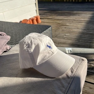 Vit keps från Polo Ralph Lauren - Snygg vit keps från Polo Ralph Lauren med den klassiska broderade loggan i blått framtill. Kepsen har justerbar rem baktill för perfekt passform. Perfekt för soliga dagar!
