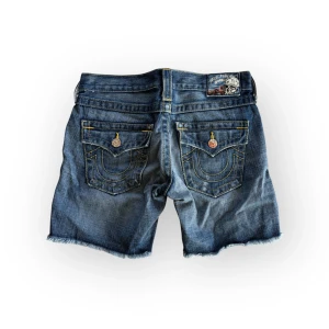 True Religion långa jeansshorts - Snygga blå jeansshorts med slitna detaljer och fransar vid bensluten. Shortsen har klassiska fem fickor och dekorativa sömmar på bakfickorna. Perfekta för sommaren. Är osäker på om det är herr eller dam modell. Storlek W25 passar S. Midja 38 längd 35