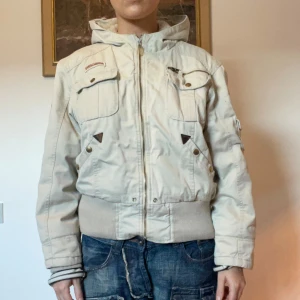 Beige jacka från Airborne - Snygg beige jacka från Airborne med dragkedja och flera fickor med knappar. Jackan har en hög krage och ribbade muddar vid ärmslut och nederkant. Rätt så varm.