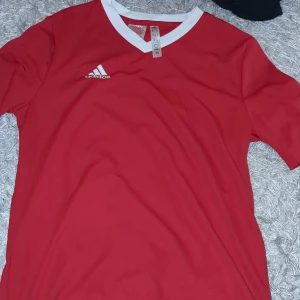 Röd fotbollströja från Adidas - Säljer en snygg röd fotbollströja från Adidas med vit V-ringning och korta ärmar. Perfekt för träning eller match. Adidas-loggan är tryckt på bröstet. Passar dig som vill ha en stilren och bekväm tröja på planen.