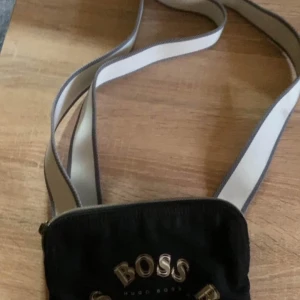 Svart axelväska från Hugo Boss - Snygg svart axelväska från Hugo Boss med ett stilrent tryck av märkesnamnet i guld. Väskan har en dragkedja och en bred axelrem i kontrasterande färger. Perfekt för att bära med sig det nödvändigaste med stil. Äldre modell denna modellen hittar du inte nån stans längre.