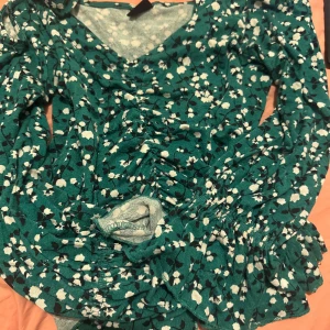 Grön blommig blus från Ginatricot - Snygg grön blus  med blommigt mönster från ginatricot. Den har en v-ringad design och långa ärmar med volangdetaljer vid ärmslutet. Perfekt för en stilfull look! Har en lite mer croppad look.