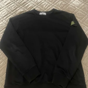 Svart tröja från Stone Island - Säljer en stilren svart tröja från Stone Island med deras ikoniska märke på ärmen. Tröjan har långa ärmar och en klassisk rund halsringning. Perfekt för en avslappnad och trendig look.