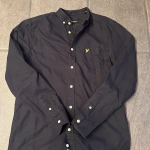 Svart skjorta från Lyle & Scott - Säljer en stilren svart skjorta från Lyle & Scott med klassisk krage och knappar framtill. Skjortan har en broderad logotyp i gult på bröstfickan och långa ärmar med knappstängning vid ärmsluten.