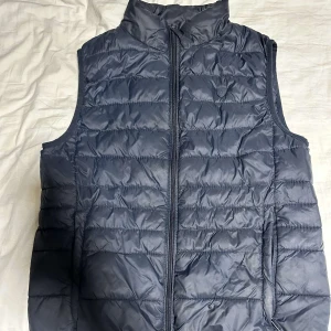 Mörkblå quiltad dunväst - Snygg mörkblå quiltad dunväst med dragkedja framtill. Perfekt för lager-på-lager under kyliga dagar. Västen har en hög krage för extra värme och stil. Från Álvaro 