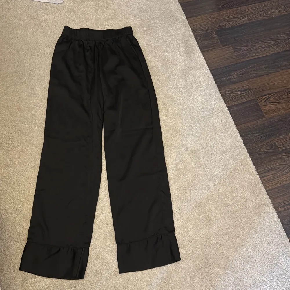 Snygga svarta silkes byxor från Gina Tricot med volangdetalj vid bensluten. Byxorna har en loose passform och är perfekta för en stilren look. Elastisk midja för extra komfort.. Farkut & Housut.