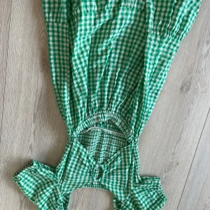 Grön- och vitrutiga klänning från Lindex - Charmig grön- och vitrutiga klänning från Lindex med smockdetaljer i midjan och korta ärmar. Perfekt för en somrig look med sin lätta och luftiga design. Klänningen har en söt knytning framtill och är tillverkad i bomull.
