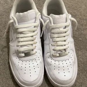 Vita Nike Air Force sneakers - Säljer ett par klassiska vita Nike Air Force sneakers. De har en stilren design med snörning och perforerade detaljer på ovansidan för extra ventilation. Perfekta för en casual look.
