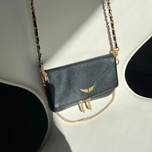 Svart zadig voltaire väska med gulddetaljer  - Elegant svart axelväska i skinn med guldkedja och dekorativt emblem på framsidan. Väskan har en praktisk dragkedja på baksidan för extra förvaring. Perfekt för att ge en stilren touch till din outfit. Dustbag medkommer😊