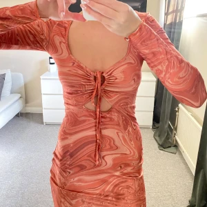 Mönstrad klänning med snörning - Trendig klänning i en härlig orange nyans med ett unikt mönster. Den har långa ärmar och en snygg snörning framtill som ger en fin detalj. Perfekt för att sticka ut!