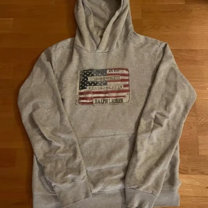 Grå hoodie från Ralph Lauren - Säljer en grå hoodie från Ralph Lauren med en stor amerikansk flagga och text på framsidan. Tröjan har en klassisk huva och ribbade muddar vid ärmslut och nederkant. Perfekt för en avslappnad stil.