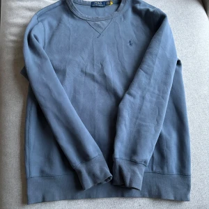 Blå tröja från Ralph Lauren - Säljer en stilren blå tröja från Ralph Lauren med rund hals och broderad logotyp på bröstet. Tröjan har långa ärmar och ribbade muddar vid ärmslut och nederkant. Perfekt för en avslappnad look. 