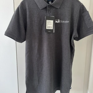 Grå pikétröja från Stakater - Säljer en stilren grå pikétröja från Stakater med broderad logga på bröstet. Tröjan har korta ärmar och en klassisk krage med knappar. Perfekt för en avslappnad men ändå snygg look.