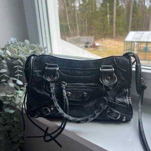 Snygg trendig väska - Snygg svart handväska i skinn med dragkedja och detaljerade spännen. Väskan har både handtag och en avtagbar axelrem. Passar till alla tillfällen 💕