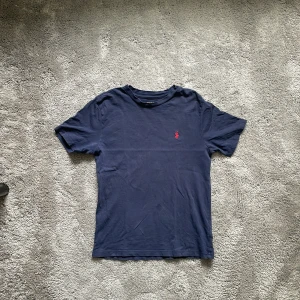 Mörkblå t-shirt från Polo Ralph Lauren - Säljer min sons mörkblå t-shirt från Polo Ralph Lauren med det klassiska röda broderade logotypen på bröstet. Perfekt för en avslappnad look. T-shirten är kortärmad och gjord i mjuk bomull.