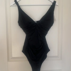 Svart baddräkt från Cubus  - Snygg svart baddräkt från Cubus  med justerbara axelband och rynkdetaljer framtill. Perfekt för en stilren look vid poolen eller stranden. Tillverkad i ett bekvämt material som formar sig efter kroppen. Är även oanvänd 