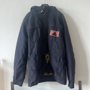 Mörkblå parkajacka från Scotch & Soda - Säljer en mörkblå parkajacka från Scotch med huva och flera praktiska fickor. Jackan har en broderad detalj på bröstet och dragsko i midjan för justerbar passform. Perfekt för kyligare dagar.