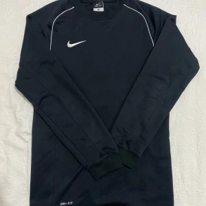 Svart långärmad tröja från Nike - Säljer en svart långärmad tröja från Nike med Dri-FIT-teknologi. Tröjan har en vit Nike-logga på bröstet och vita detaljer vid axlarna. Perfekt för träning eller vardagsbruk.