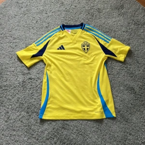 Sverige fotbollströja  - Säljer en gul fotbollströja från Adidas med Sveriges landslagsemblem på bröstet. Tröjan har korta ärmar och blå detaljer, inklusive ränder på axlarna och sidopaneler. Perfekt för fotbollsfans! 
