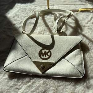 Vit kuvertväska från Michael Kors - Elegant vit kuvertväska från Michael Kors med ett stilrent design och logotypdetalj i metall. Väskan har en justerbar axelrem och en triangulär flik framtill. Perfekt för att bära det nödvändigaste med stil.