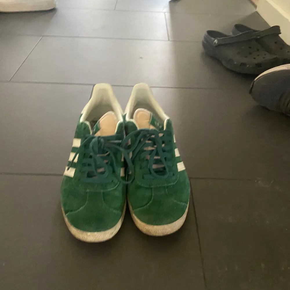 Snygga gröna Adidas Gazelle sneakers i mocka med vita detaljer och snörning. Perfekta för en stilren och sportig look. De ikoniska tre ränderna och Gazelle-loggan i guld ger en klassisk touch. Man får även med box och ett par vita sko snören så man kan ha det man gillar mest. Kengät.