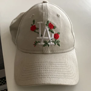 Beige keps från New Era med rosbroderi - Snygg beige keps från New Era med broderade röda rosor och vit 'LA'-logga framtill. Justerbar rem baktill för perfekt passform. Perfekt för att ge din outfit en unik touch.