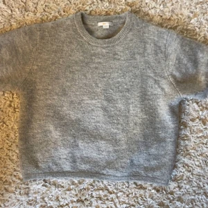 Grå stickad tröja från Gina Tricot - Säljer en mysig grå stickad tröja från Gina Tricot. Tröjan har en rund halsringning och korta ärmar, perfekt för lager-på-lager-stil. Den är enkel och stilren, vilket gör den lätt att matcha med olika outfits. 