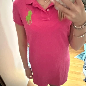 Rosa klänning från Ralph Lauren - Säljer en snygg rosa klänning från Ralph Lauren med en grön logga på bröstet. Klänningen har korta ärmar och en klassisk krage. Perfekt för en avslappnad stil med en touch av elegans.