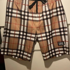 Burrberry badshorts - Snygga badshorts från Burberry med klassiskt rutigt mönster i beige, svart, vit och röd. De har en bekväm passform med snörning i midjan för justerbarhet. Perfekta för sommarens stranddagar. Sitter perfekt på mig, är 182. 
