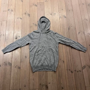 Grå merinoull hoodie - Säljer en stilren grå hoodie med långa ärmar och justerbar huva. Perfekt för en avslappnad look. Den har en klassisk passform och är gjord i ett mjukt material. OBS! ena metall grejen på högra sidan av tråden som används för att spänna luvan saknas.