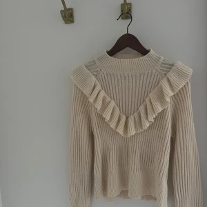 Beige stickad tröja med volangdetalj - Säljer en stilren beige stickad tröja med volangdetalj runt halsen. Tröjan har långa ärmar och en ribbad struktur. 💞
