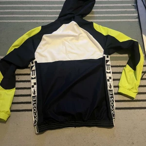 Flerfärgad vindjacka från Lacoste - Säljer en snygg vindjacka från Lacoste i vitt, svart och neongult. Jackan har en dragkedja framtill och en huva. Den har långa ärmar och elastiska muddar. Perfekt för en sportig look! STORLEK M
