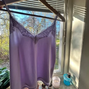 Ljuslila linnetopp med spets - Säljer en ljuslila linnetopp med tunna axelband och vacker spetsdetalj längs urringningen. Toppen har en mjuk och glansig yta som ger en lyxig känsla. Perfekt att styla till olika outfits.
