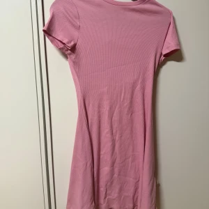 Rosa ribbad kortklänning - Säljer en söt rosa ribbad kortklänning med rund hals och korta ärmar. Klänningen har en figurnära passform och är perfekt för varma dagar. Enkel och stilren design som passar till många tillfällen.